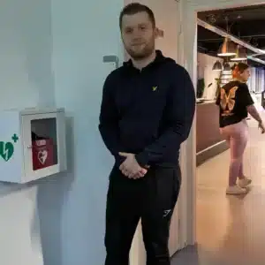 Man standing beside a red AED mounted on a wall in an indoor setting.