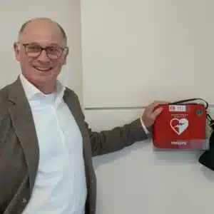 Smiling man with glasses pointing at a red wall-mounted AED.