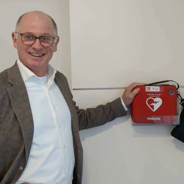 Smiling man in blazer pointing to a red defibrillator mounted on an indoor wall.