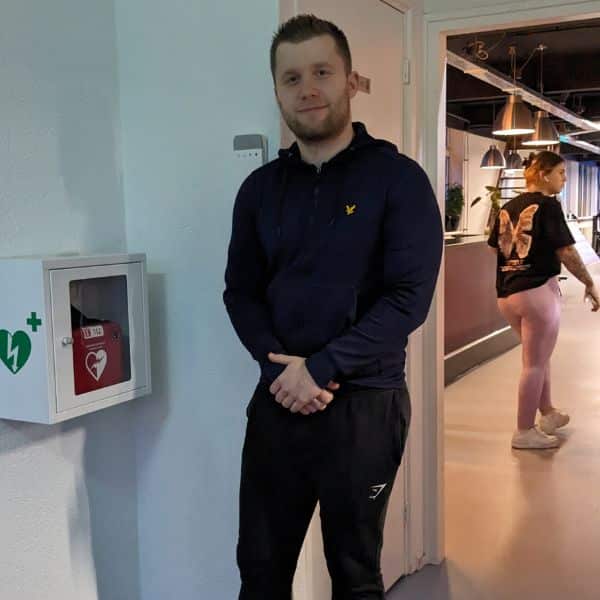 Man standing near a defibrillator in a gym hallway with people training in background.