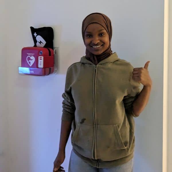 Smiling woman giving a thumbs up next to a wall-mounted defibrillator indoors.