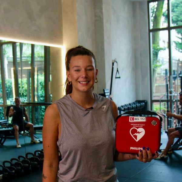 Woman in a gym holding a red defibrillator with fitness equipment in the background.