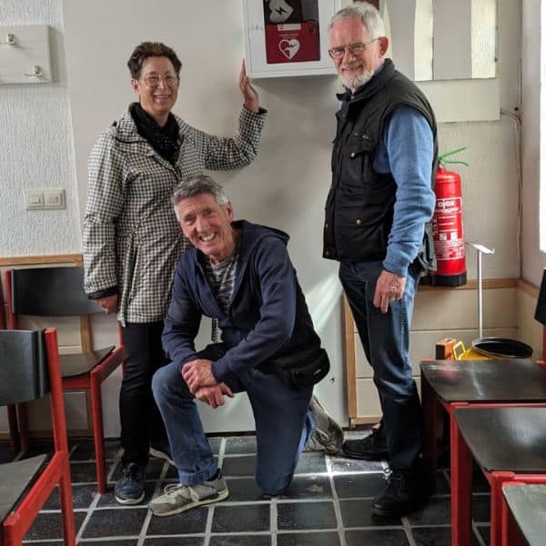 Three adults standing by a wall-mounted AED unit in a shared kitchen or cafeteria setting.