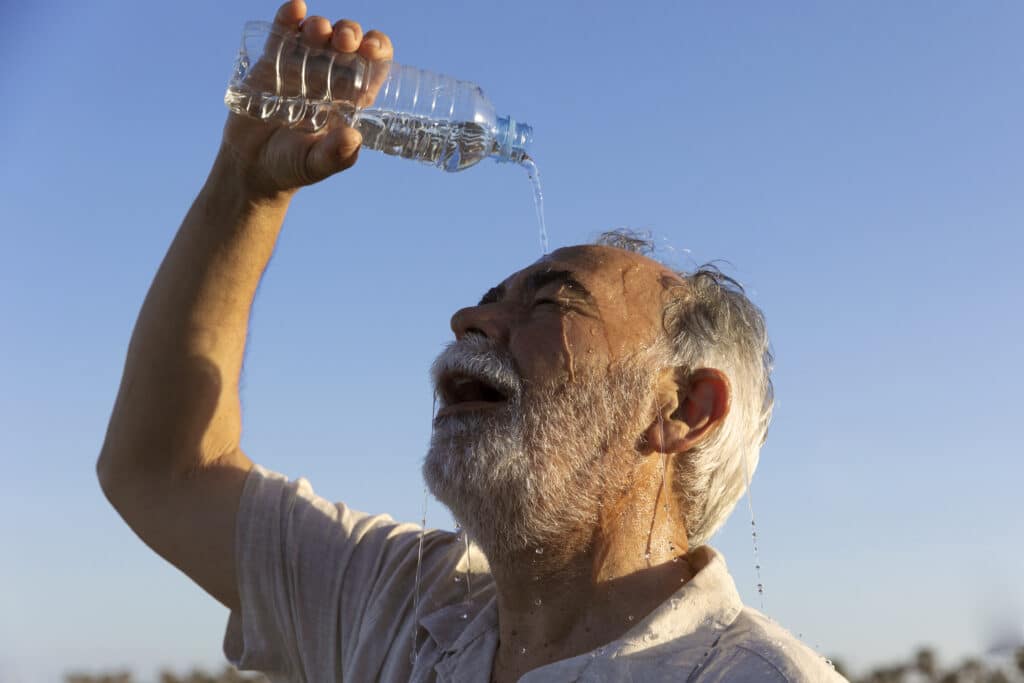 An older man pours water from a bottle over his face to cool down on a hot day under a clear blue sky.